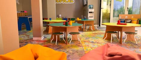 Una sala de juegos infantil con mesas y sillas de colores, un dispensador de agua y un letrero de 'Salida Exit'.