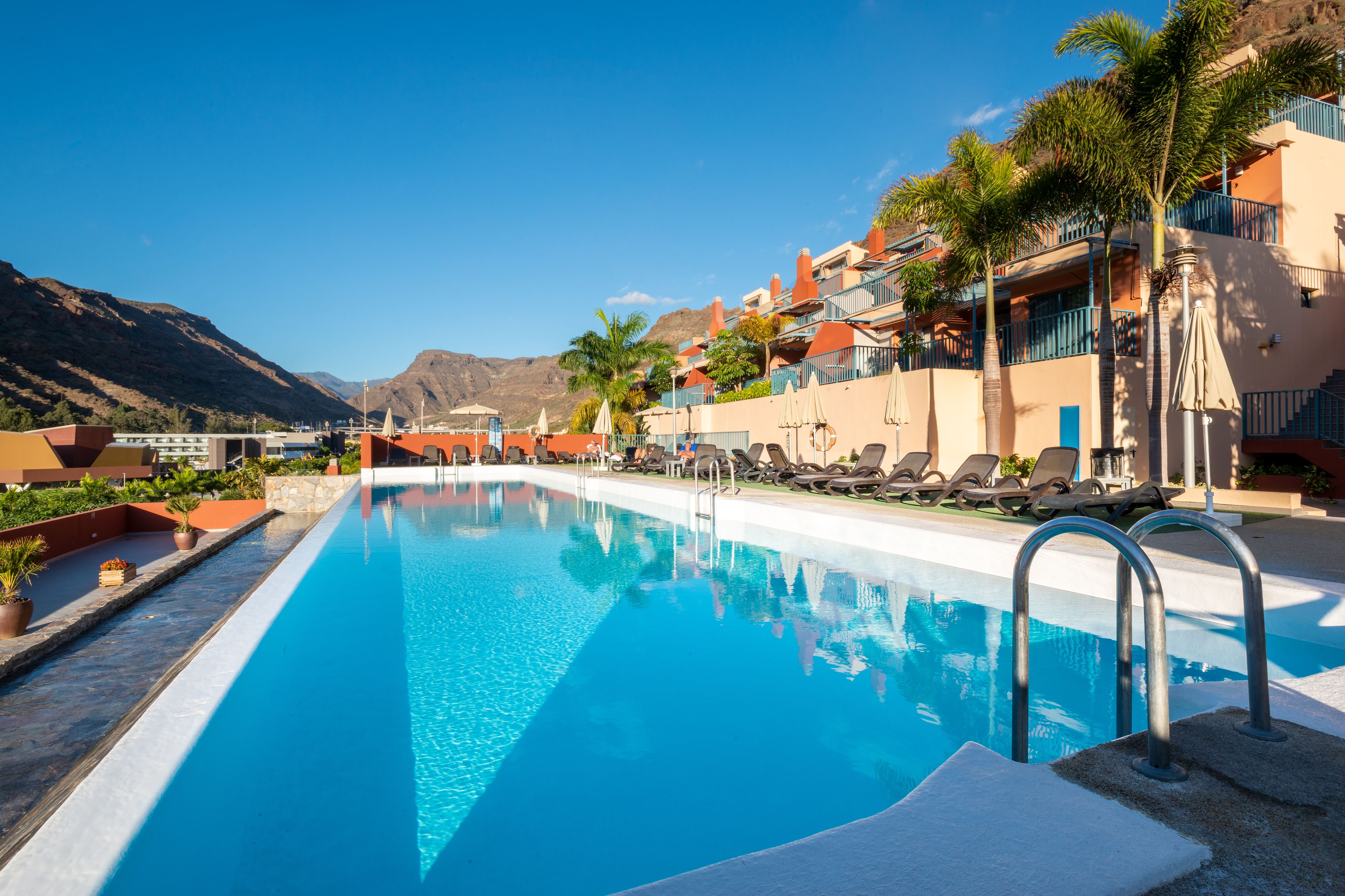 Piscina con sillas de descanso y sombrillas, rodeada de edificios con balcones y palmeras, con montañas al fondo.
