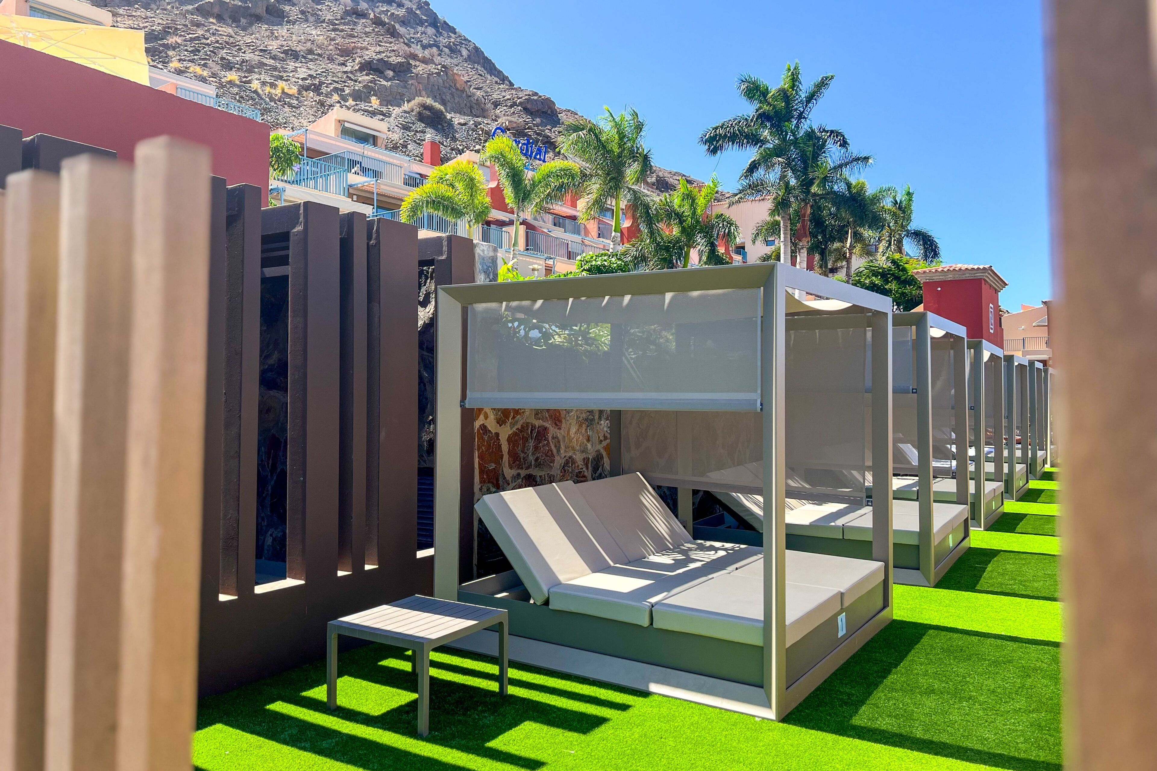 A row of sunbeds with a stone wall backdrop, a small table, and green artificial grass.