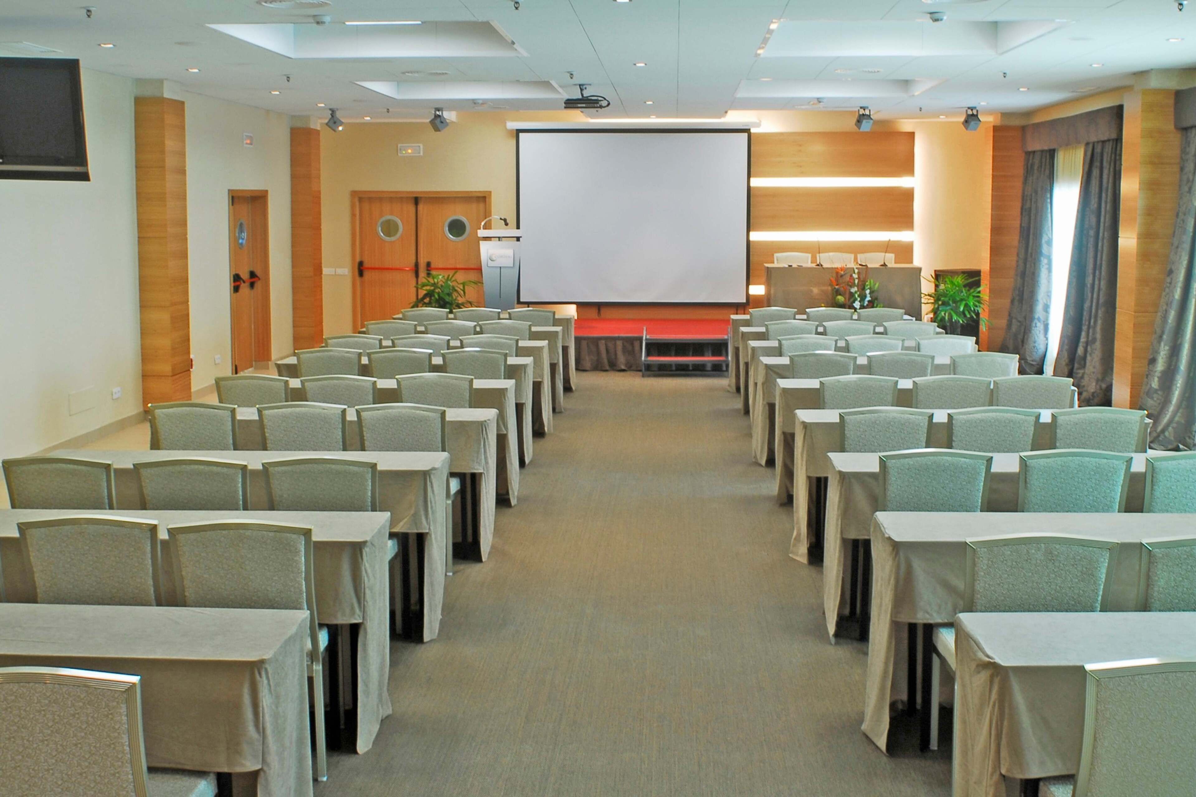 Una sala conferenze con file di tavoli e sedie, uno schermo grande in primo piano e un proiettore.