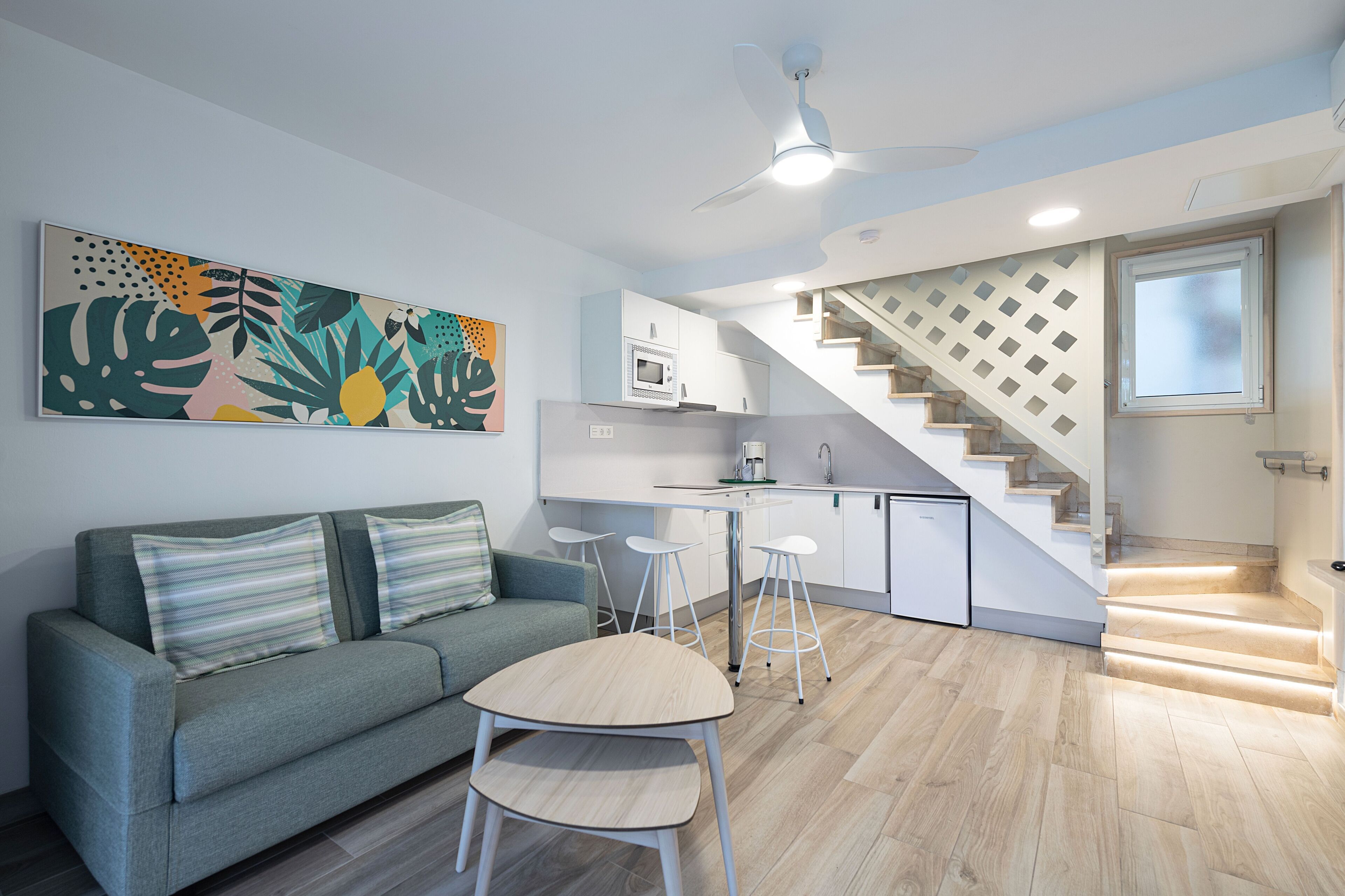 Una sala de estar moderna con un sofá gris, una mesita redonda y una cocina con armarios blancos y un microondas.