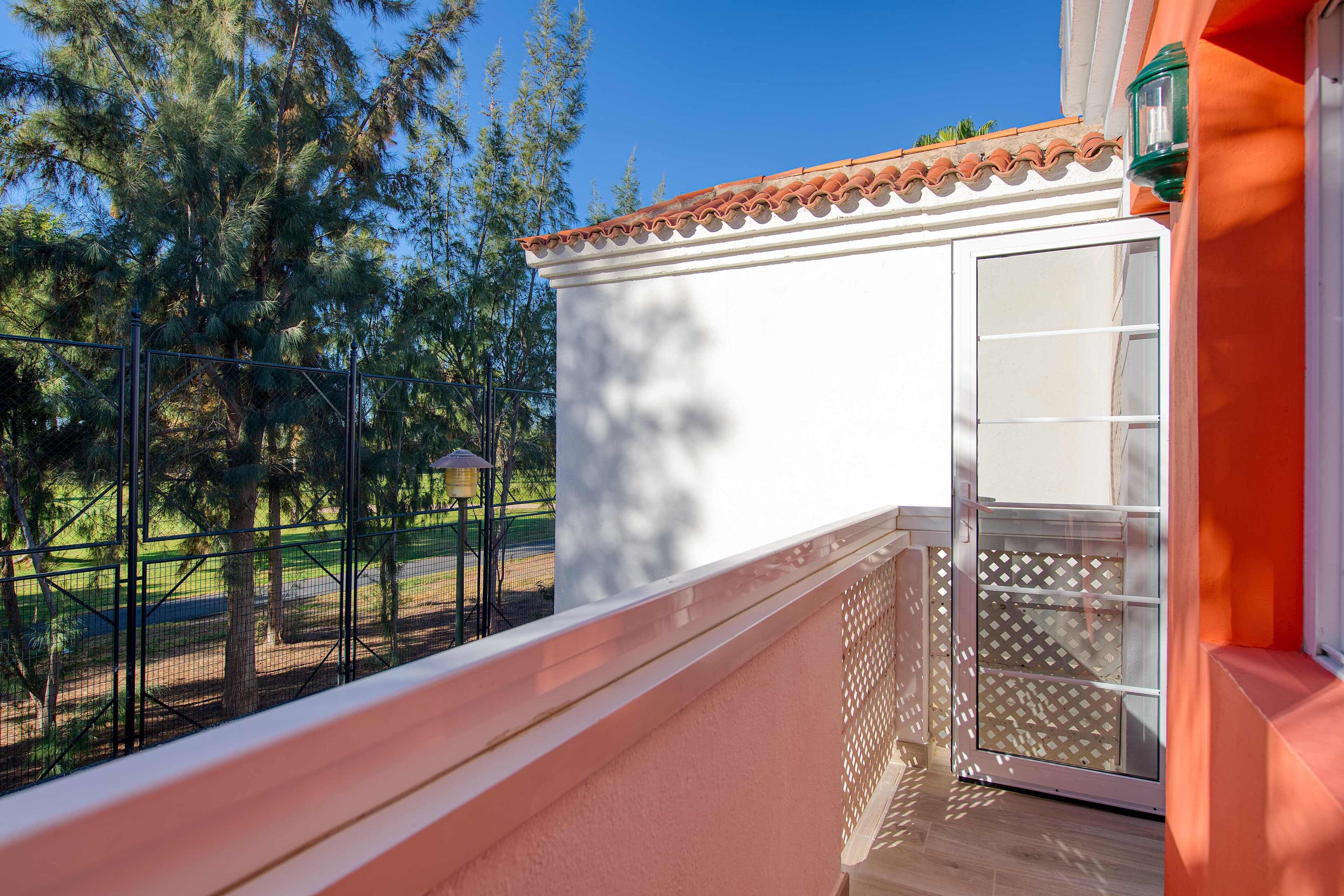 Un balcón con un muro blanco, una puerta con panel de vidrio y vista a un área exterior cercada con árboles.
