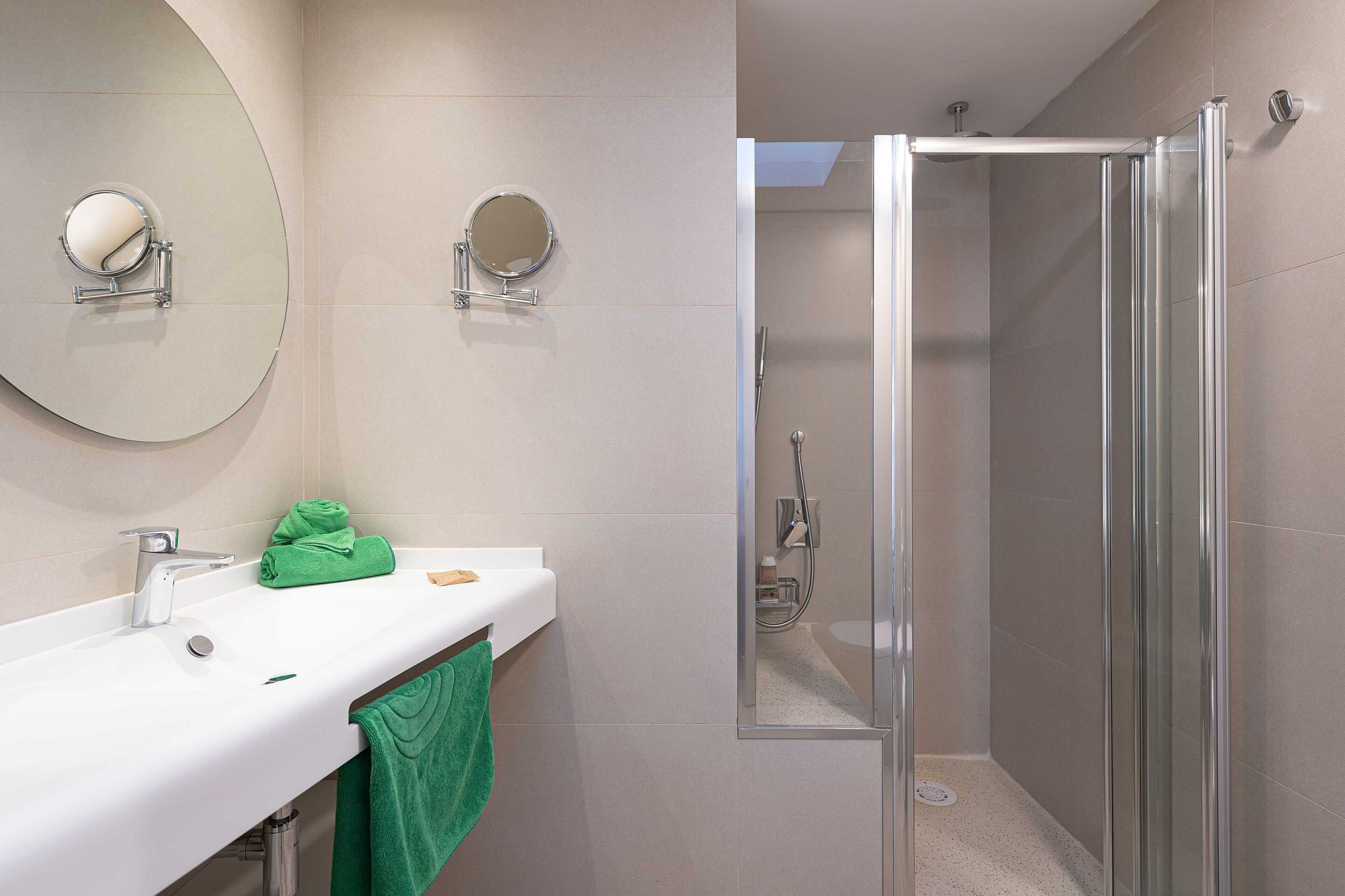 Un baño moderno con un lavamanos blanco, dos espejos redondos, una toalla verde y una ducha con mampara de vidrio.