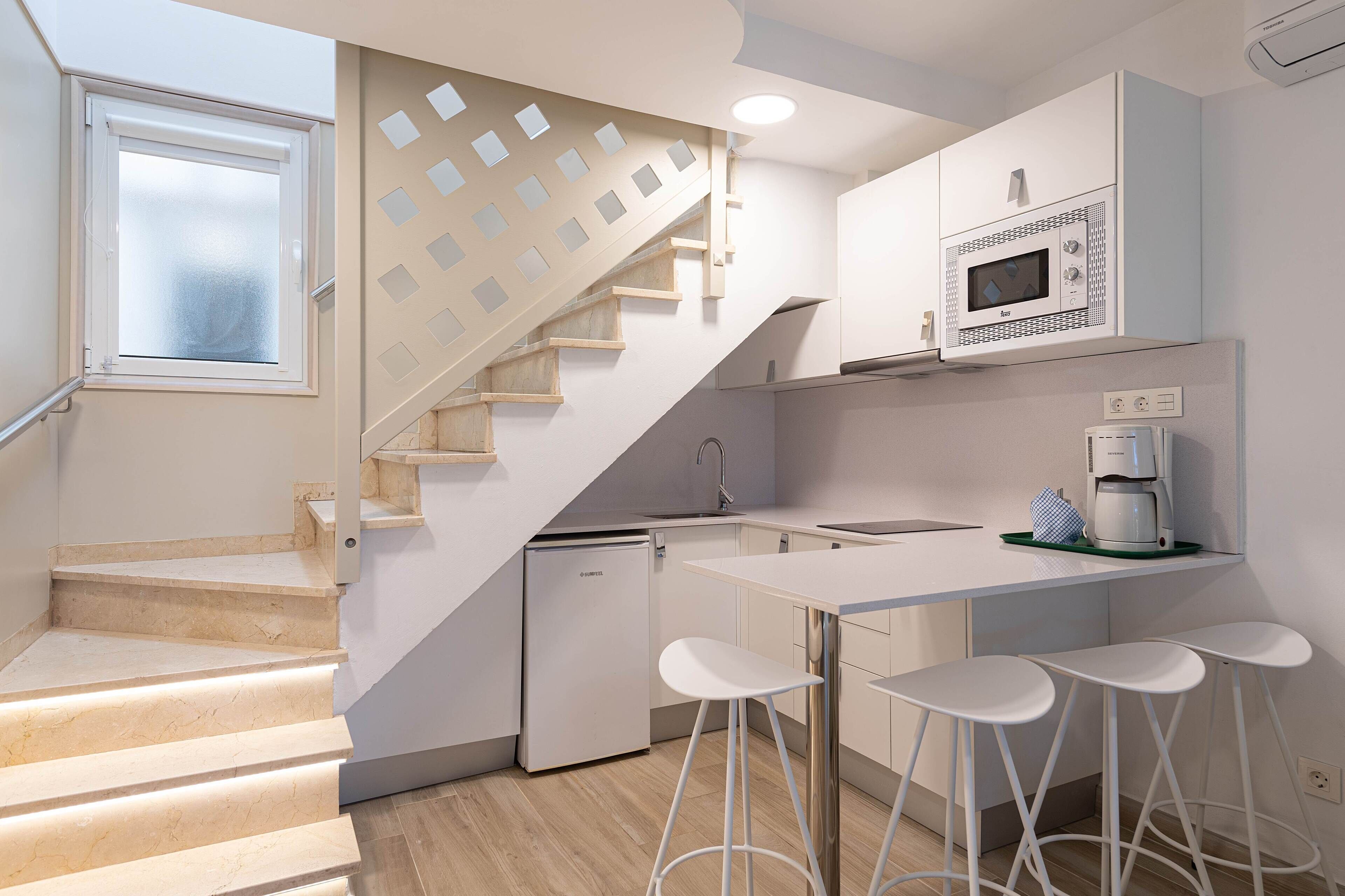 Una cocina moderna con microondas integrado, cafetera y refrigerador pequeño. Hay una isla blanca con taburetes de bar y una escalera con diseño geométrico.