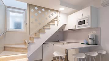 Una cocina moderna con microondas integrado, cafetera y refrigerador pequeño. Hay una isla blanca con taburetes de bar y una escalera con diseño geométrico.