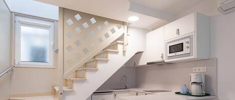 A modern kitchen with a built-in microwave, a coffee maker, and a small refrigerator. There is a white island with bar stools and a staircase with a geometric design.