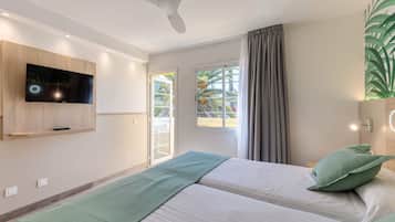Habitación de hotel moderna con televisión de pantalla plana en la pared, ventilador de techo, cama con ropa de cama verde y vista a palmeras a través de la ventana.