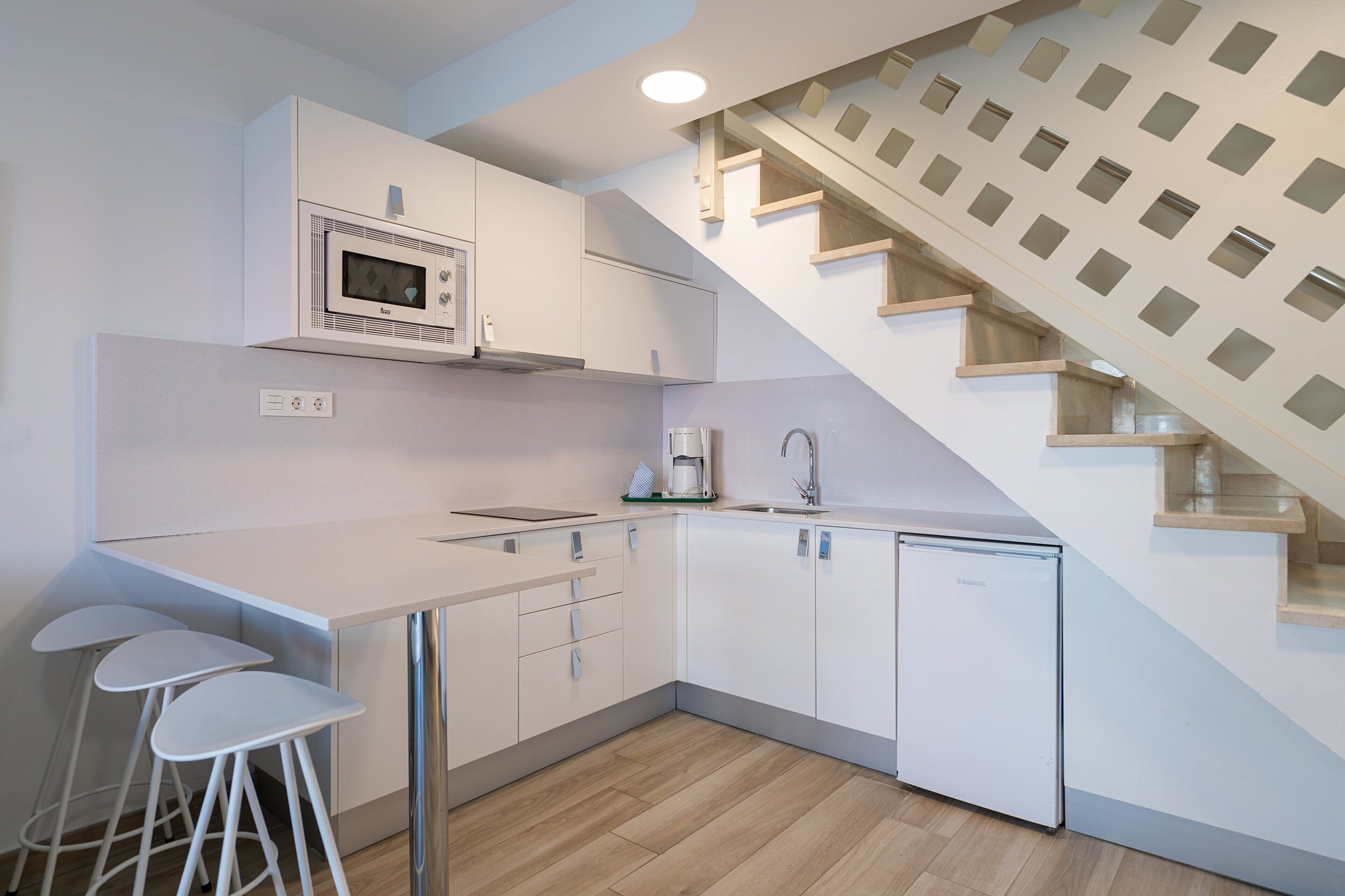 A modern kitchen with white cabinets, a microwave, a coffee maker, and a sink. There are three white bar stools and a wooden staircase with a geometric design.