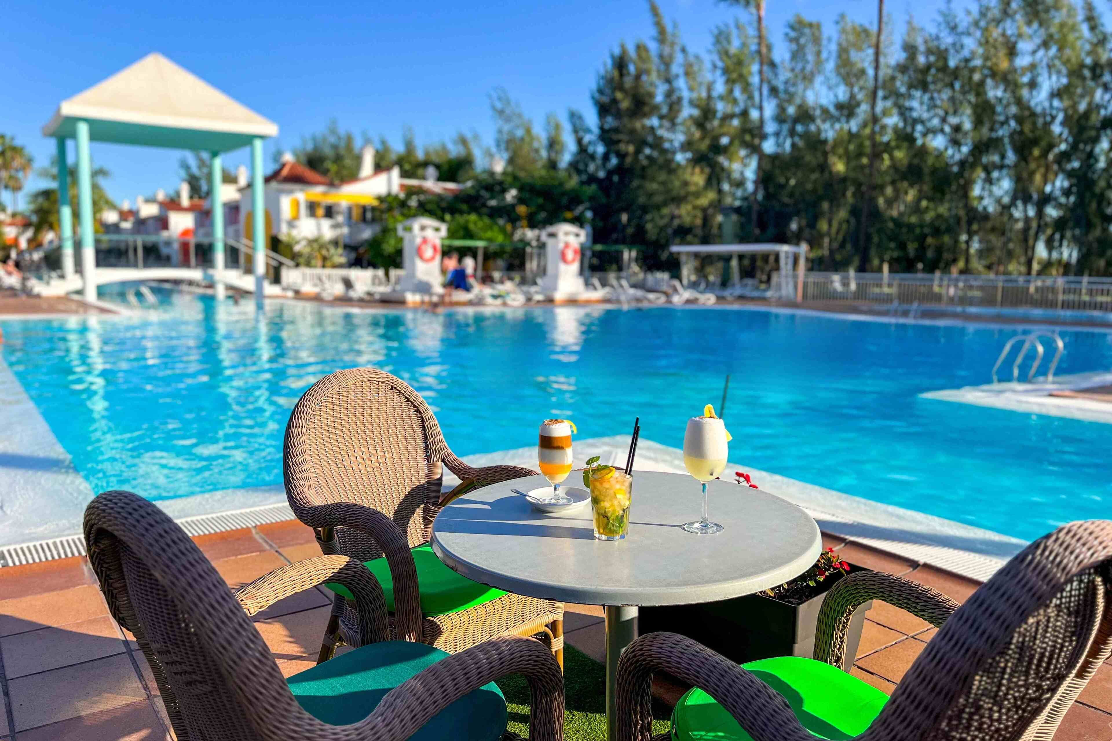 Zona de piscina con sillas de mimbre y una mesa, una bebida y una torre de salvavidas al fondo.