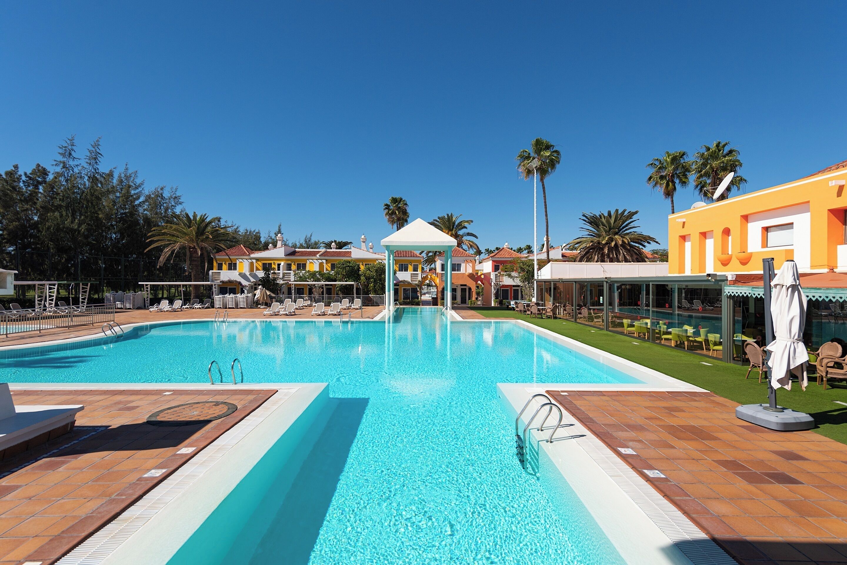 Piscina com água azul cristalina, rodeada por espreguiçadeiras e guarda-sóis, e um pavilhão no centro.