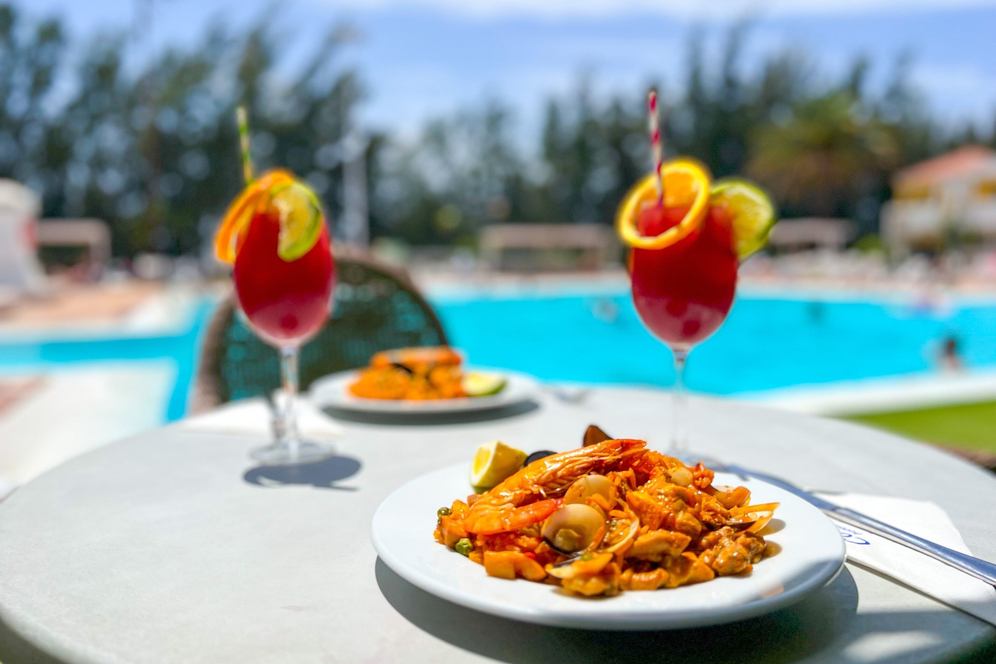 A dining table with a plate of seafood paella, two cocktails with citrus garnishes, and a pool in the background.