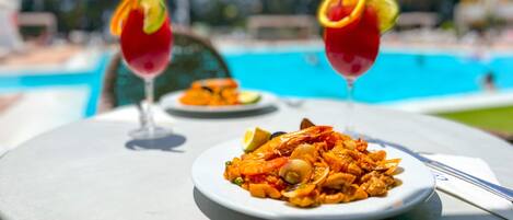 A dining table with a plate of seafood paella, two cocktails with citrus garnishes, and a pool in the background.