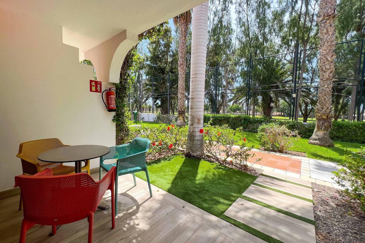 Uma área de pátio com mesa e cadeiras, um extintor de incêndio e vista para um jardim com árvores e um caminho.
