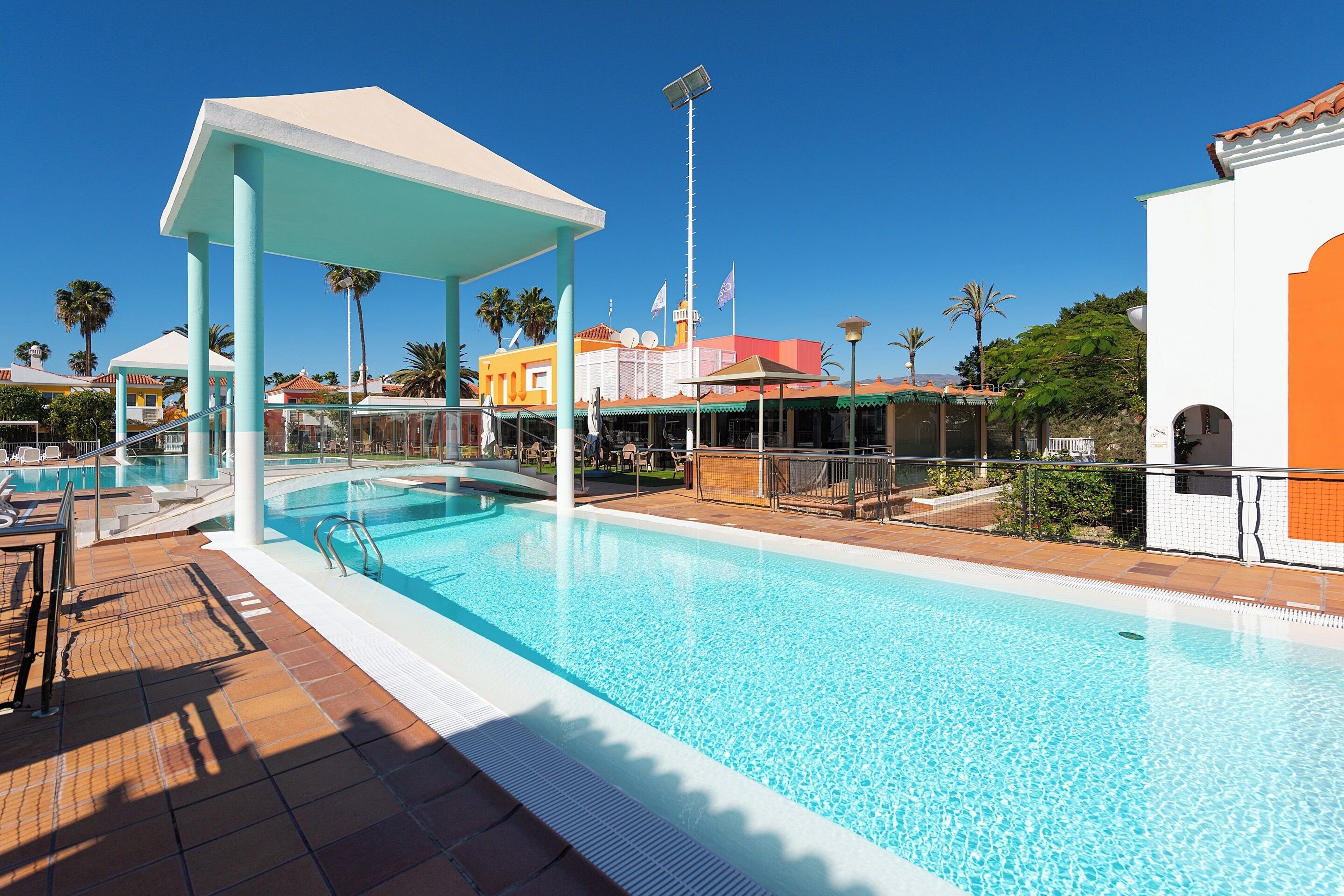 Ein Swimmingpool mit überdachtem Bereich, Palmen und Gebäuden im Hintergrund.