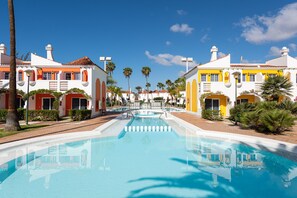 A pool area with a clear view of the pool, surrounded by buildings with red and yellow facades, palm trees, and a clear blue sky. - Bungalows Cordial Green Golf (San Bartolomé de Tirajana)