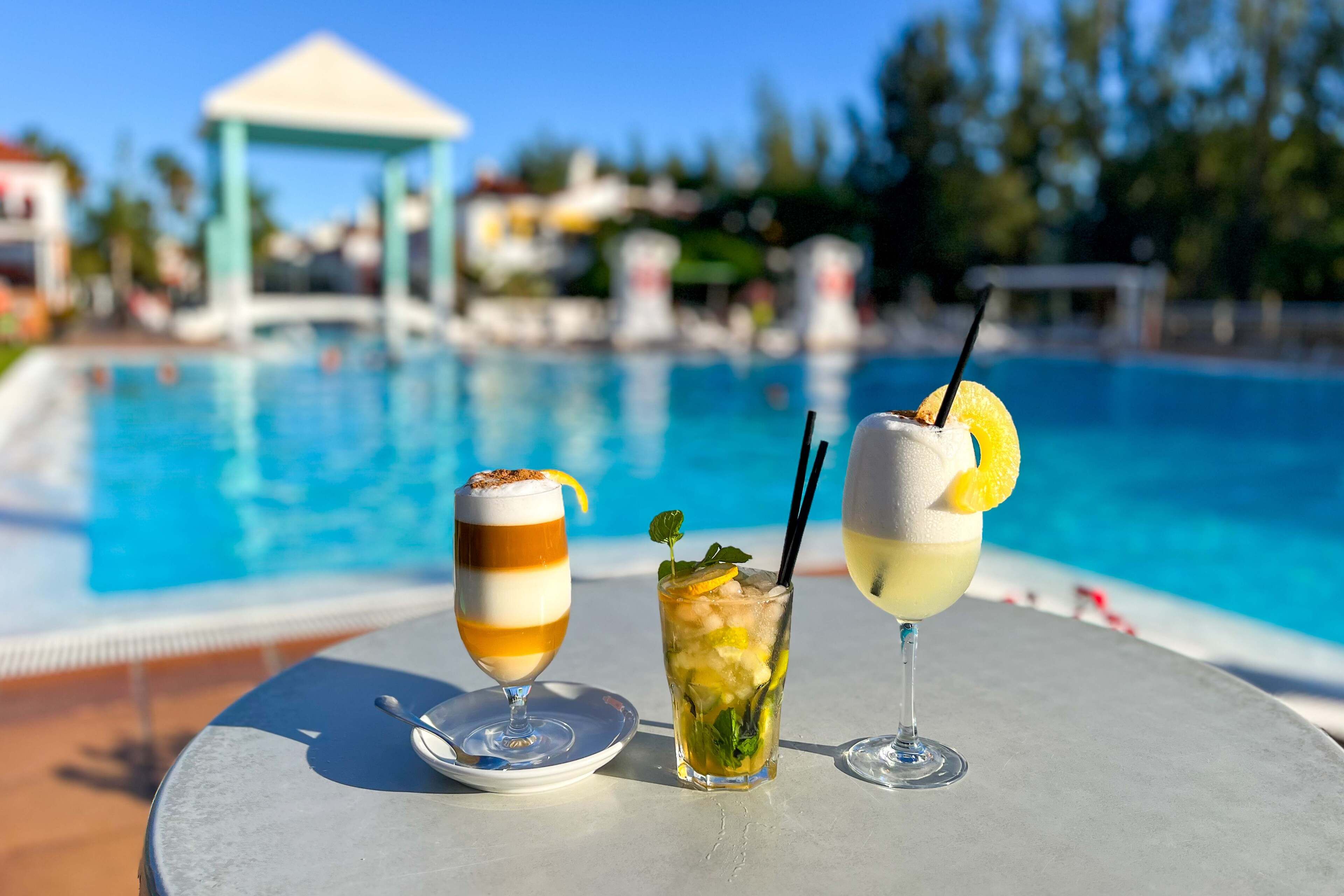 A poolside table with three drinks, a clear pool, and a gazebo in the background.