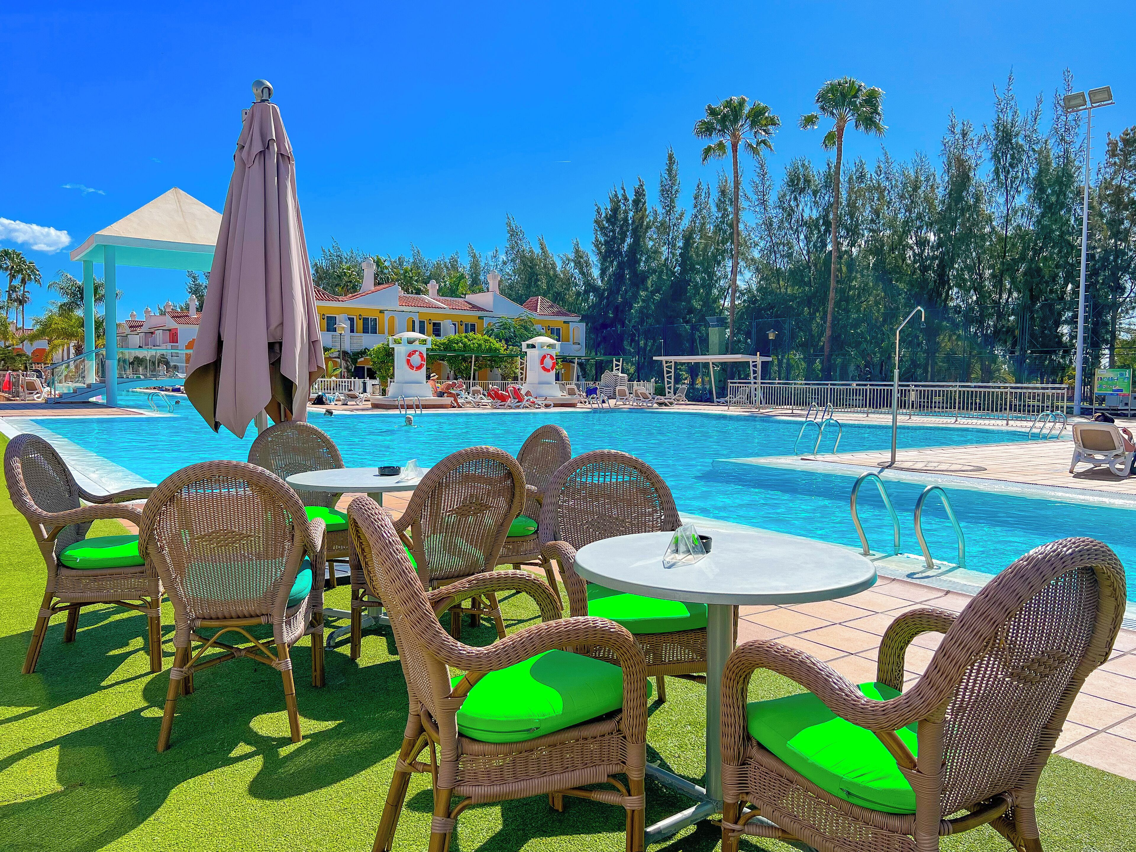 A poolside area with wicker chairs and a table, a clear blue pool, and a building with a yellow facade in the background.