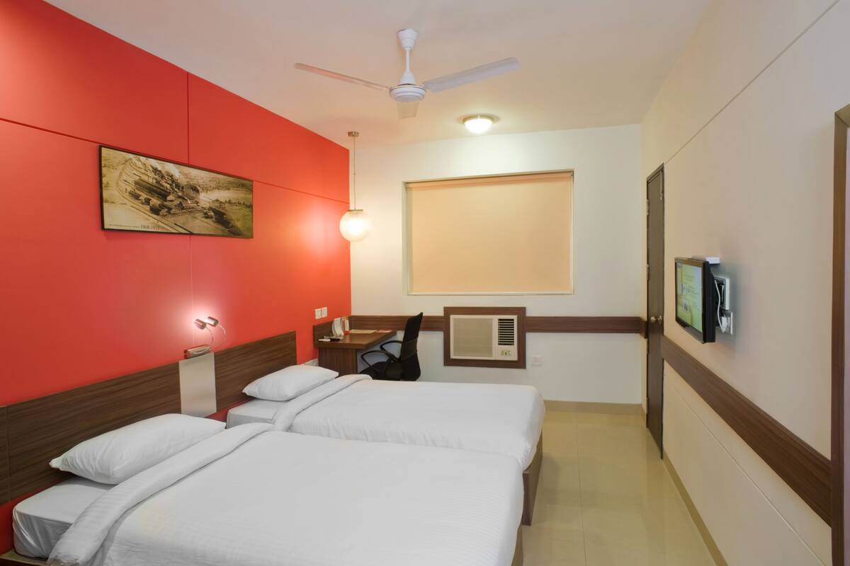 A hotel room with two beds, a ceiling fan, a mounted television, and a window with blinds.