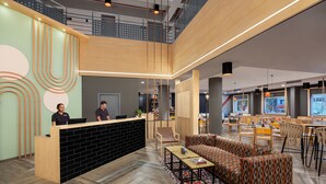 A modern reception area with a wooden reception desk, a seating area with patterned sofas, and a dining space with wooden tables and chairs.