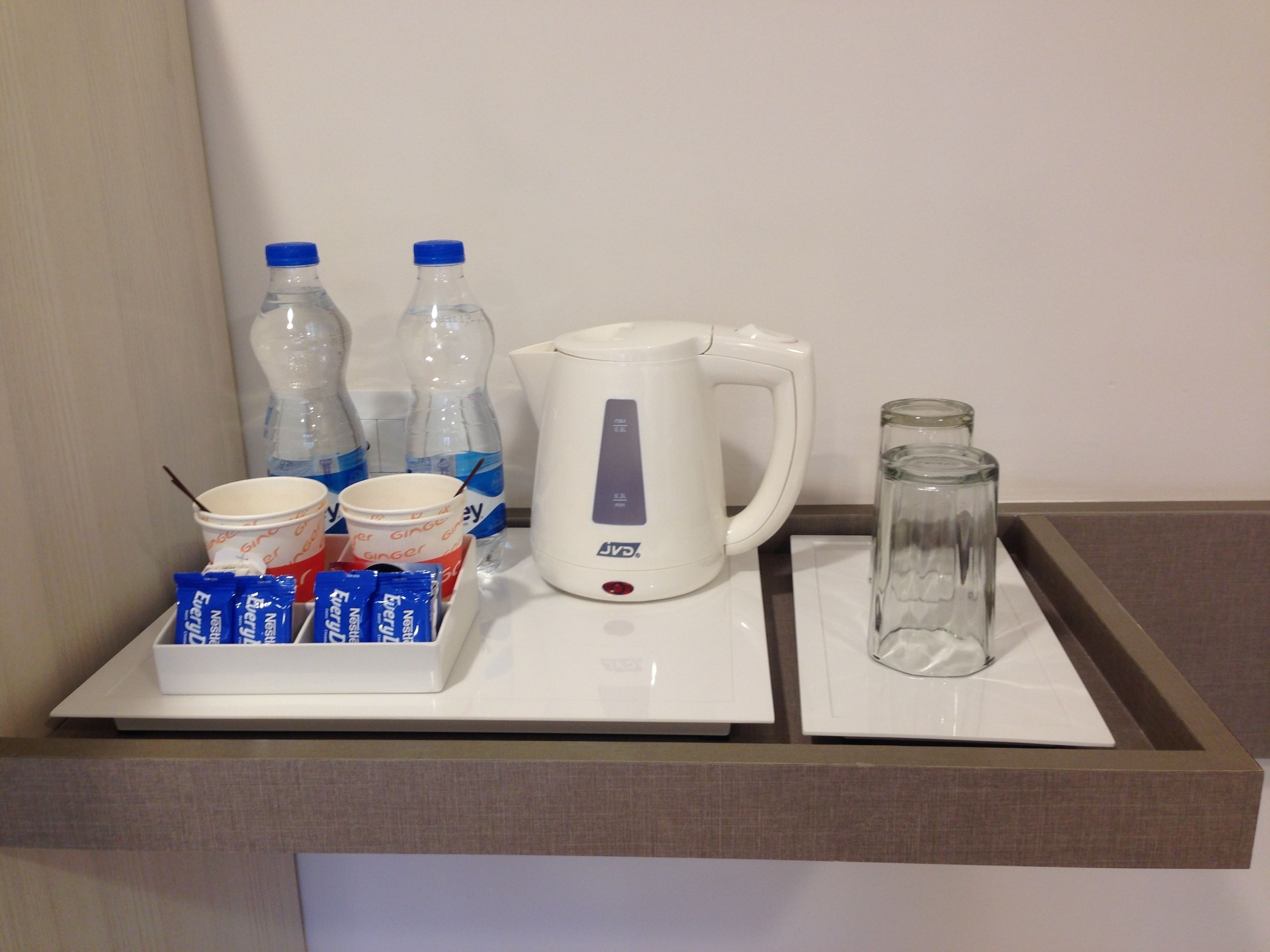Il minibar di una camera d'albergo con un bollitore bianco, due bottiglie d'acqua, un boccale di vetro e un vassoio con bustine di tè e caffè Nescafé.