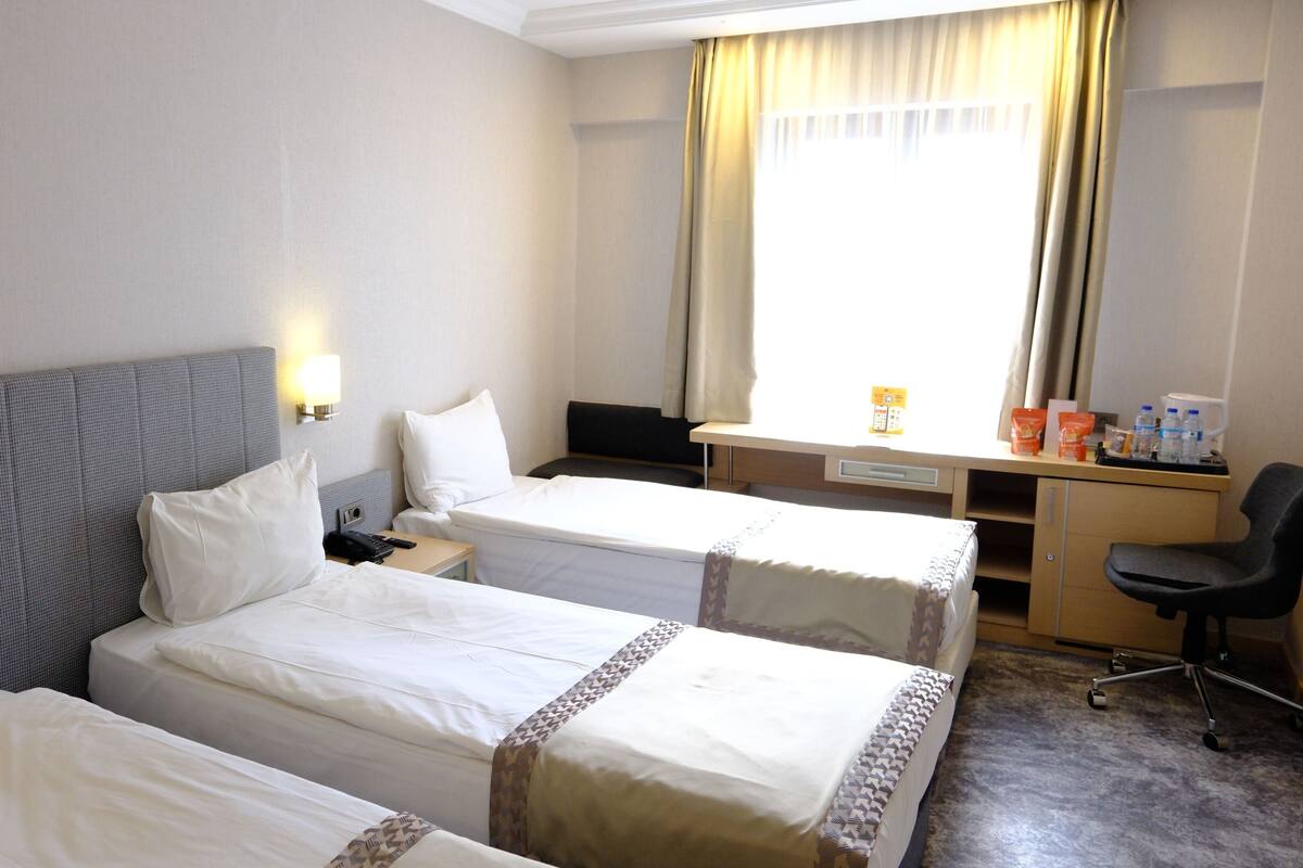 Une chambre d’hôtel avec deux lits, un bureau, une chaise, une table de chevet et une fenêtre avec des rideaux.