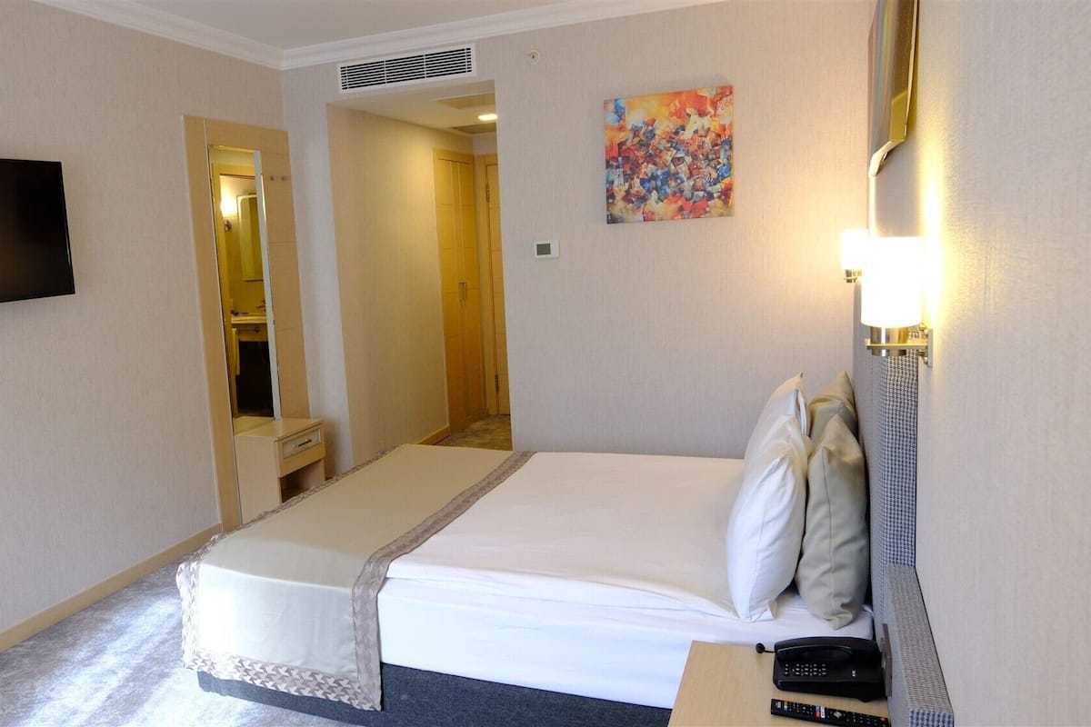 A hotel room with a bed, a TV, a painting, and a bedside table with a lamp.