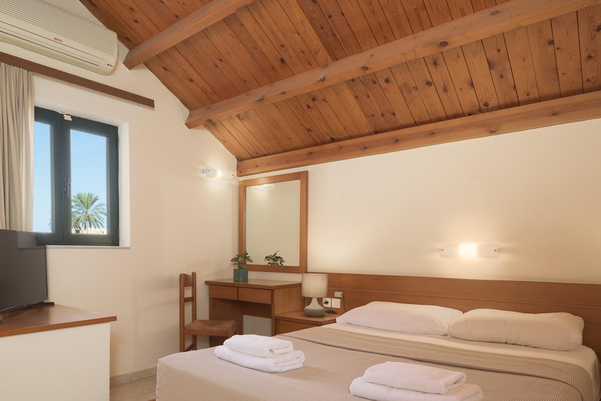 Een hotelkamer met een houten plafond, een bed met witte lakens, een bureau met stoel, een televisie en een raam met gordijnen.
