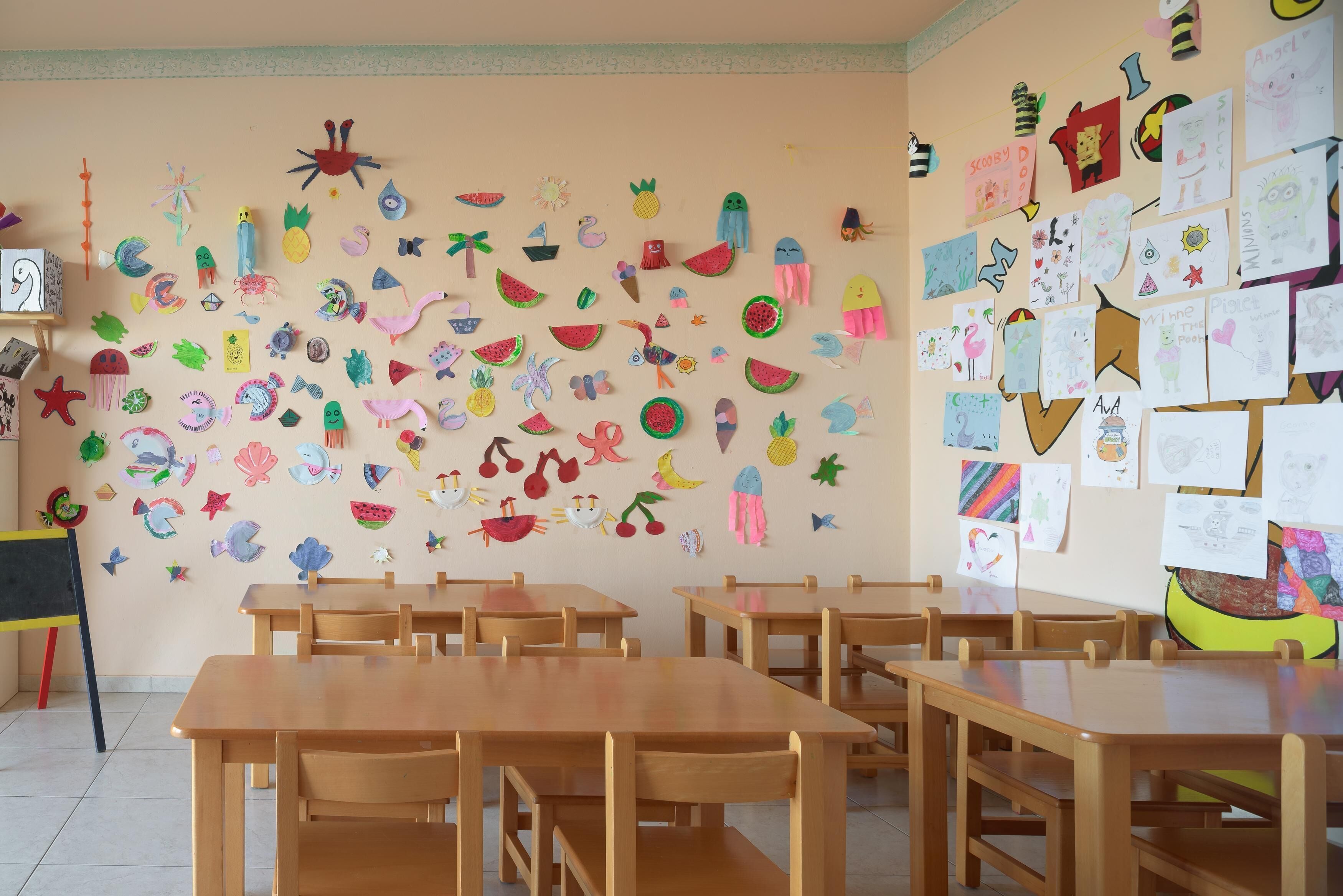 Un aula con pupitres y sillas de madera, paredes decoradas con trabajos infantiles y material educativo.