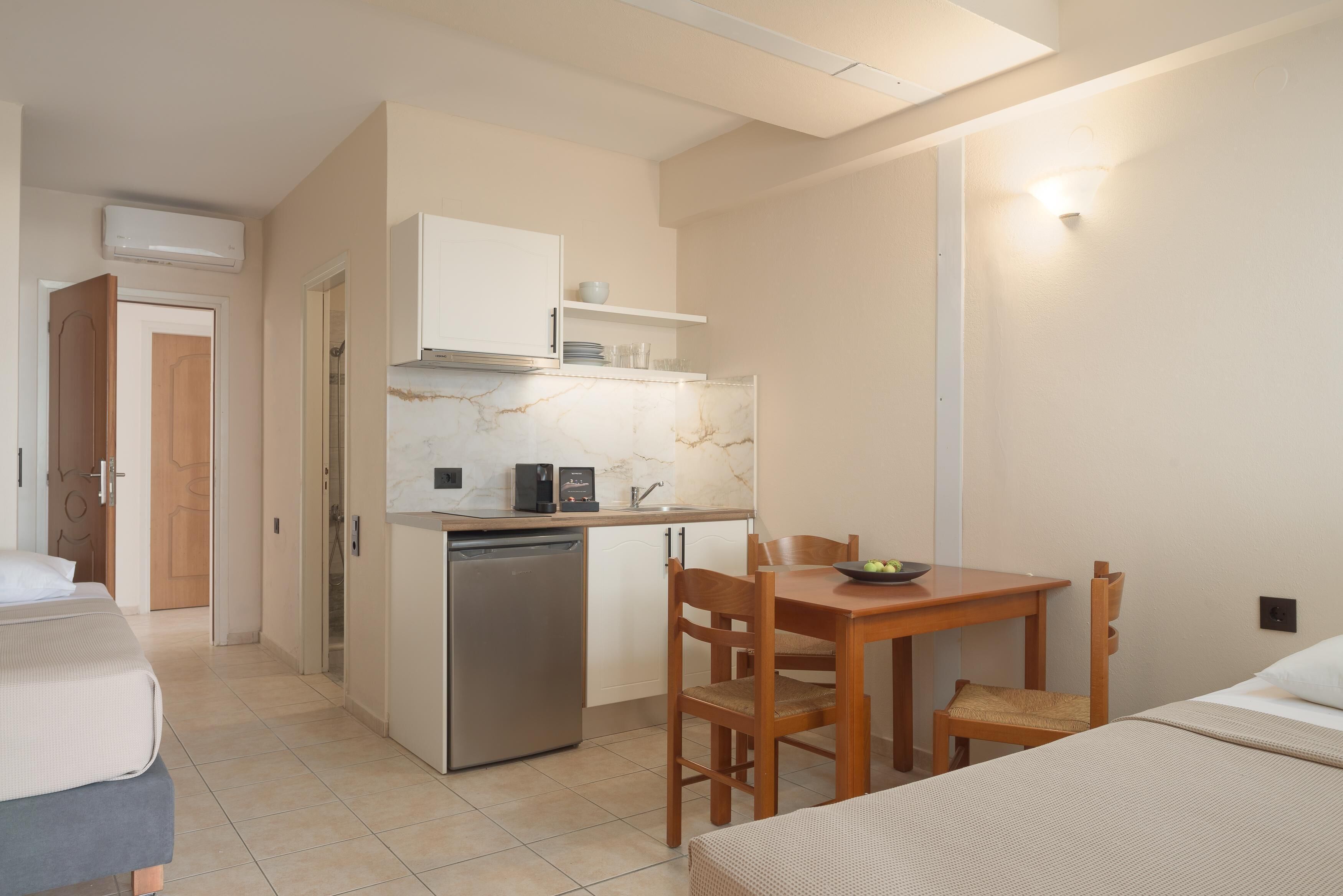 A compact hotel room with a kitchenette, a bed, a dining area with a table and chairs, and a small refrigerator.