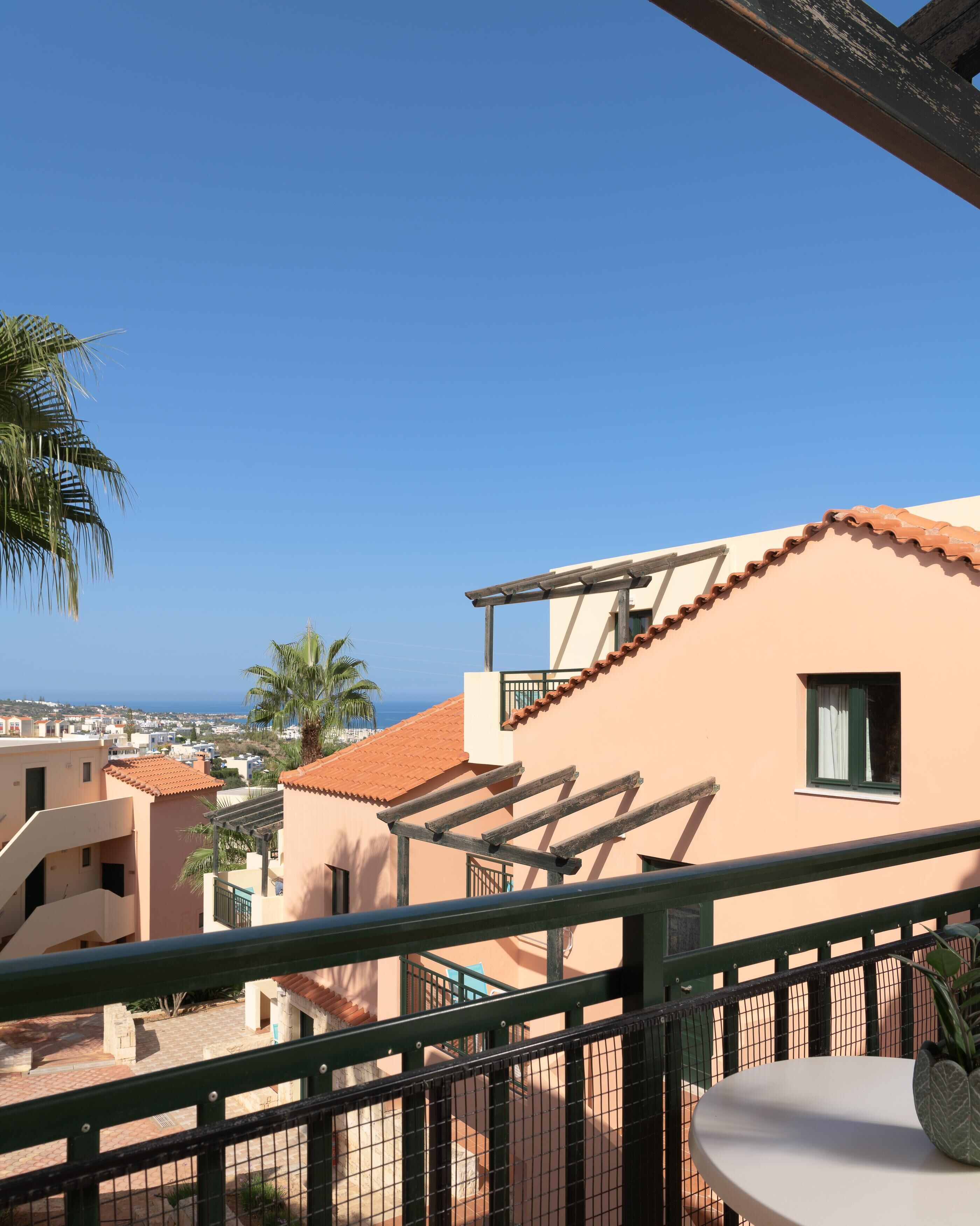 A balcony with a view of the sea, a palm tree, and neighboring buildings.