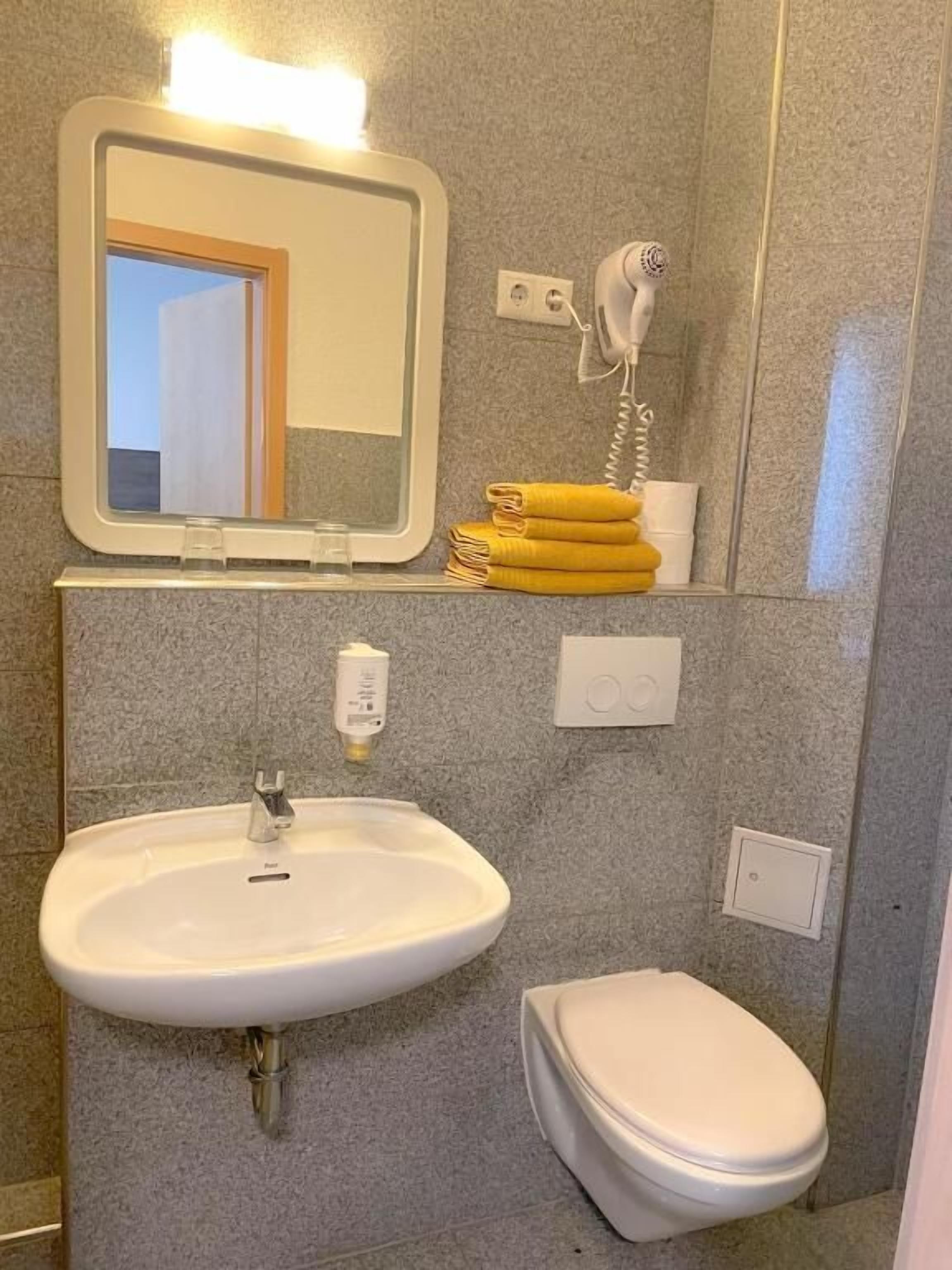 A bathroom with a sink, toilet, and a mirror with a light above it. There are towels and a hairdryer on the wall.