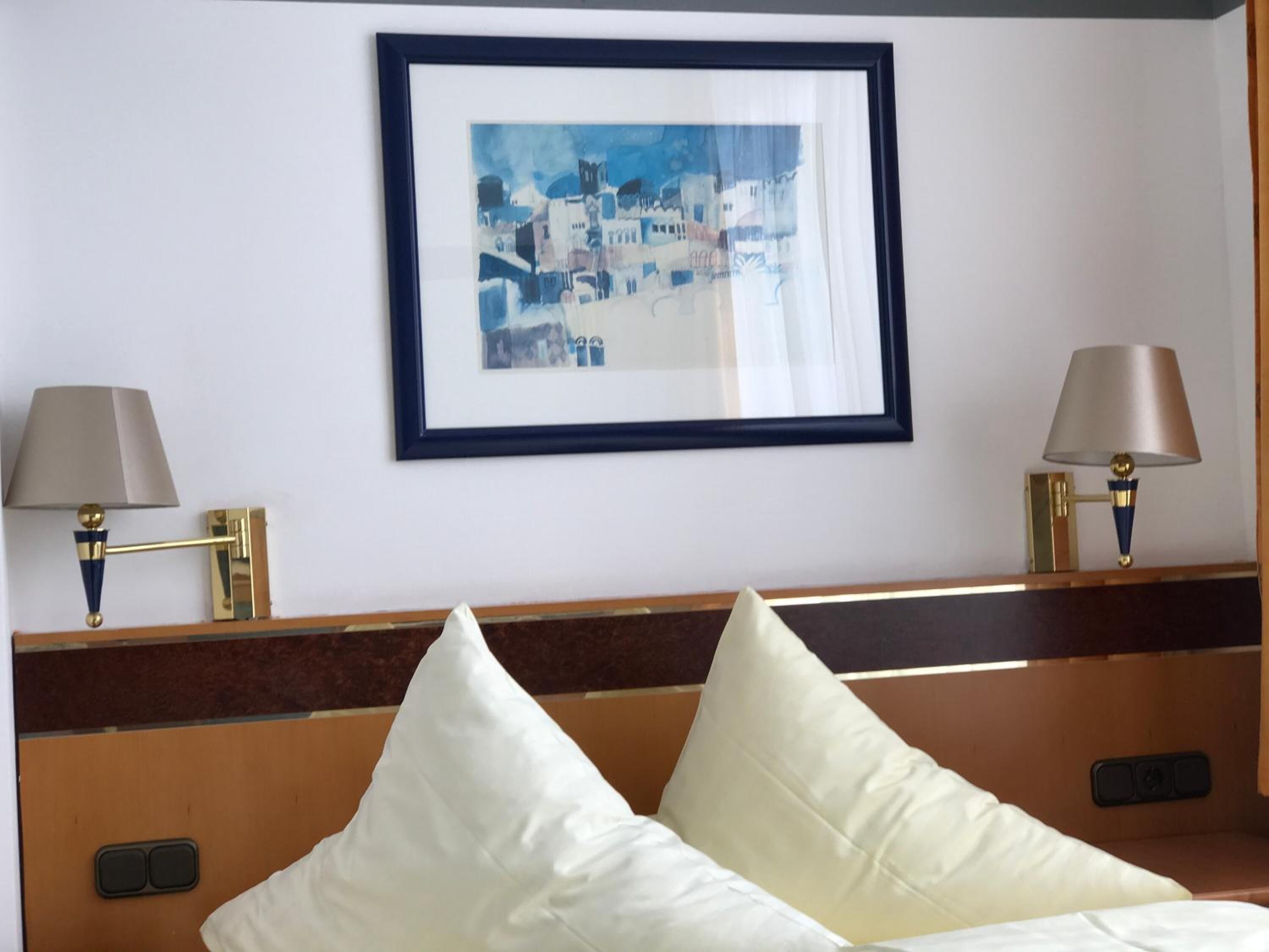 Habitación de hotel con cama, dos lámparas de noche, un cuadro en la pared y cabecera de madera.