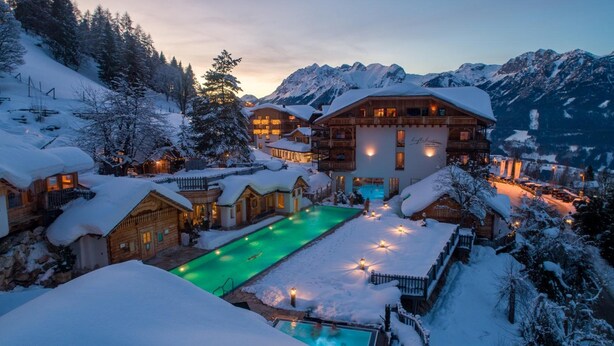 A winter resort with a pool, wooden cabins, and snow-covered mountains.