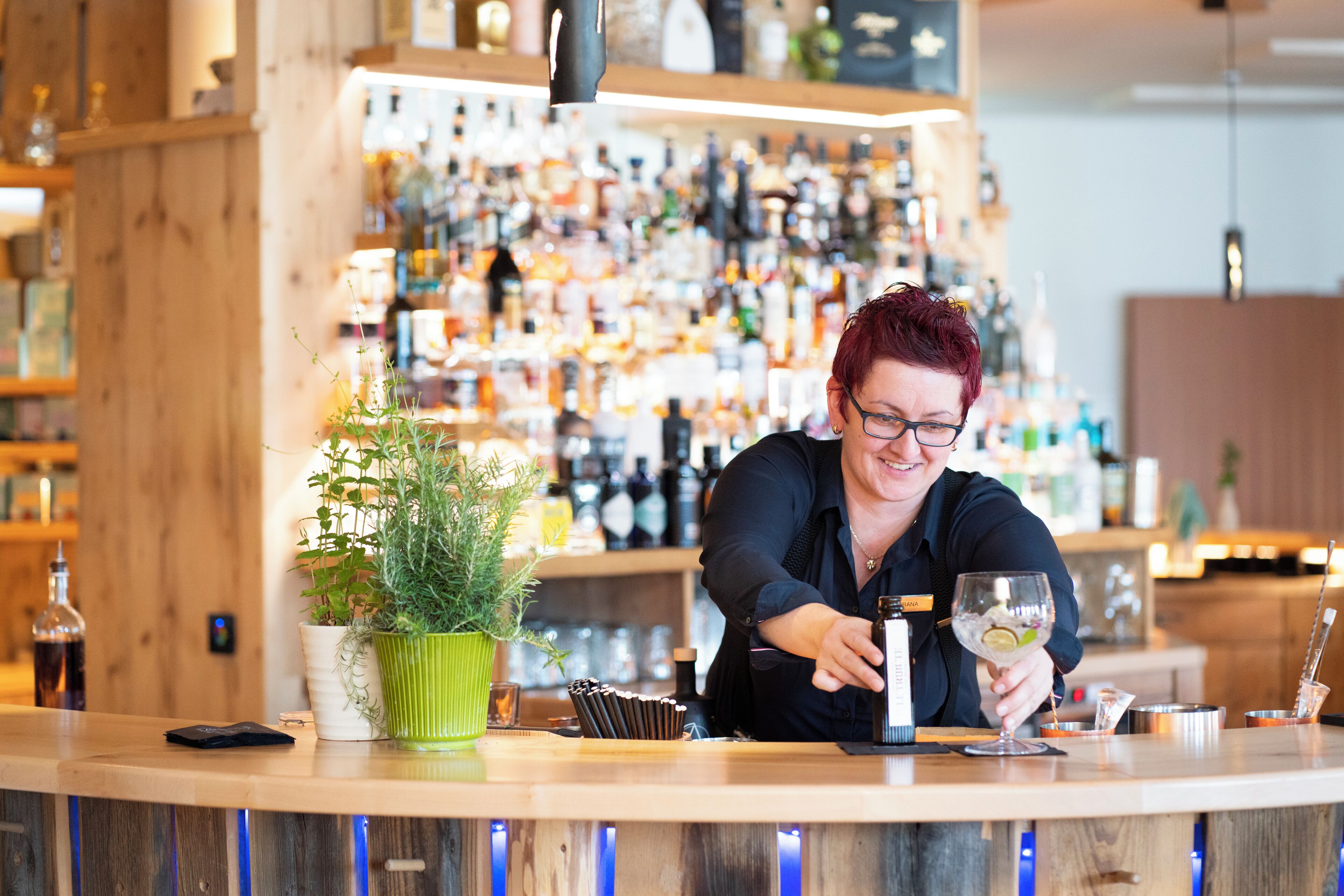 Un espace bar avec un barman préparant une boisson, des étagères remplies de bouteilles et une plante en pot.