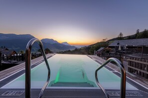Une piscine entourée de rampes métalliques, bordée d’une terrasse en bois et de bâtiments, avec une vue sur la montagne au coucher du soleil.