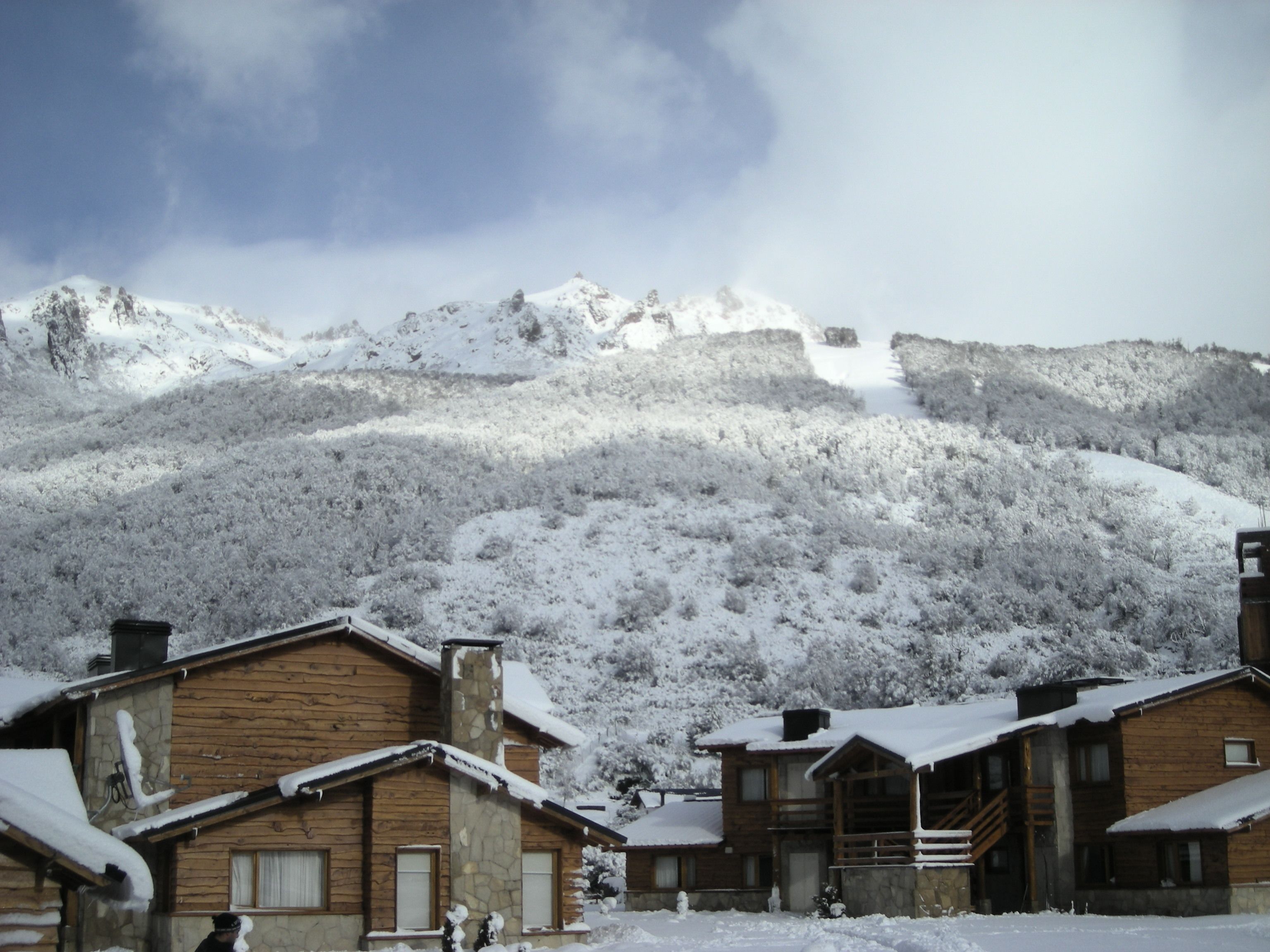 Una cabaña de madera con chimenea de piedra, rodeada de árboles cubiertos de nieve y una montaña al fondo.