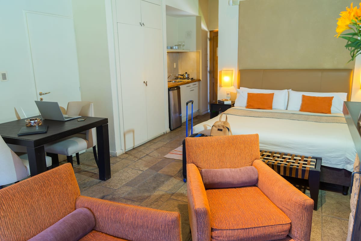 A hotel room with a bed, a desk with a laptop, two chairs, a small kitchenette, and a bathroom.