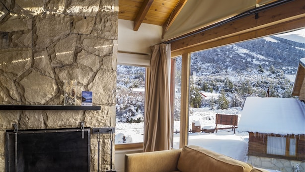 A stone fireplace, a sofa, and a view of a snowy mountain landscape through large windows. - Galileo Boutique Hotel (San Carlos de Bariloche)