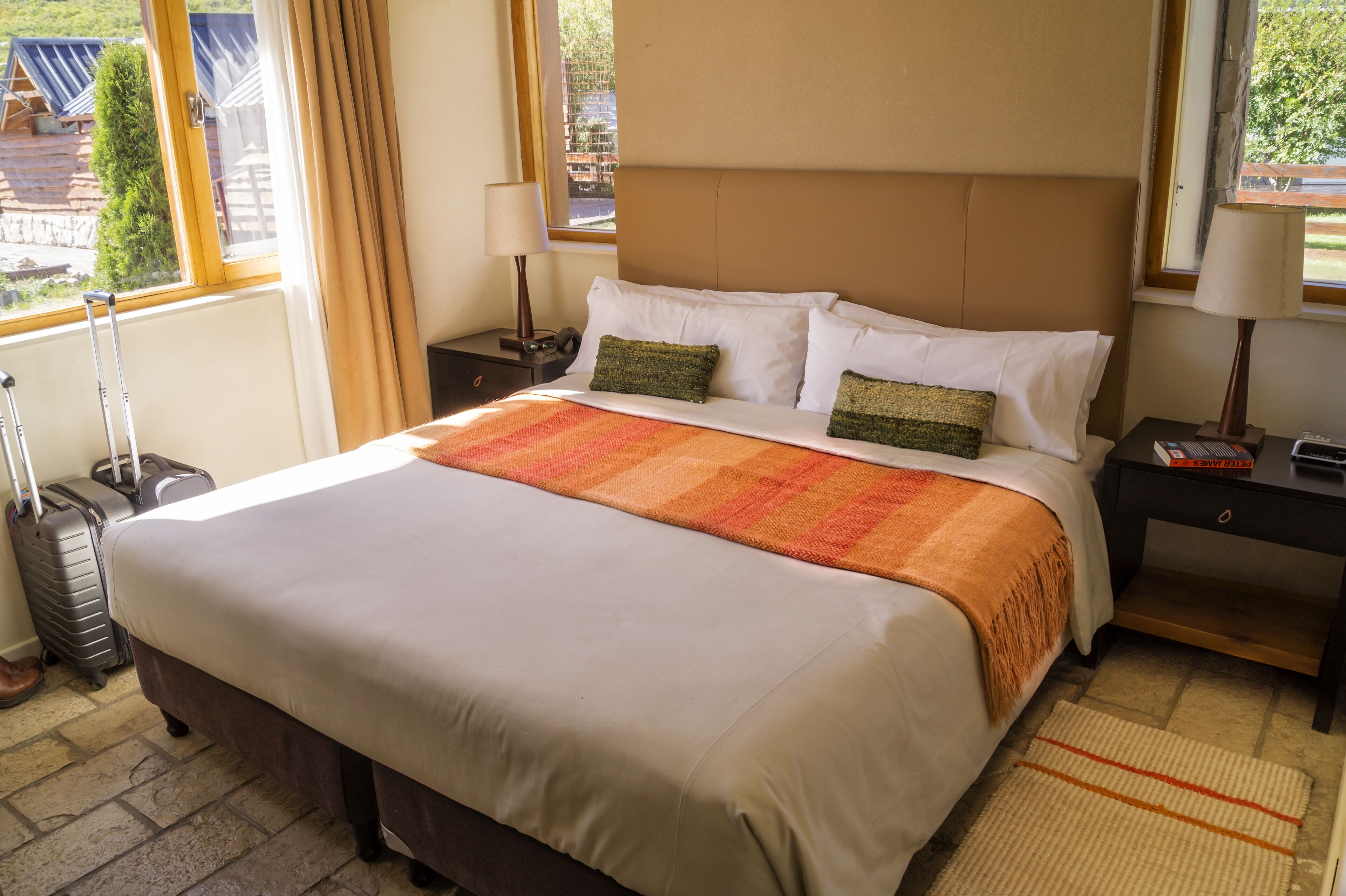A hotel room with a large bed, two bedside tables, a lamp, a suitcase, and a view of the outside.