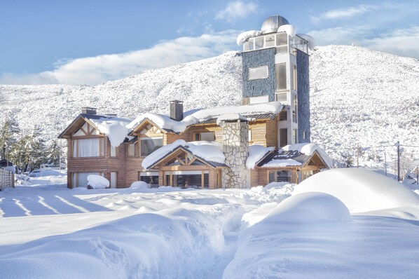 Eine schneebedeckte Lodge mit Steinaußenwand und Holzverkleidung, vor einer verschneiten Berglandschaft.