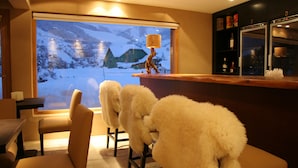A bar area with a view of snowy mountains through a large window. There are plush white chairs and a wooden counter with a decorative lamp. - Galileo Boutique Hotel (San Carlos de Bariloche)