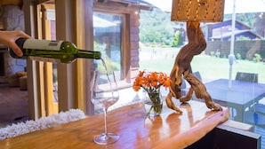 A bottle being poured into a wine glass on a wooden table with a vase of flowers and a wooden sculpture. - Galileo Boutique Hotel (San Carlos de Bariloche)