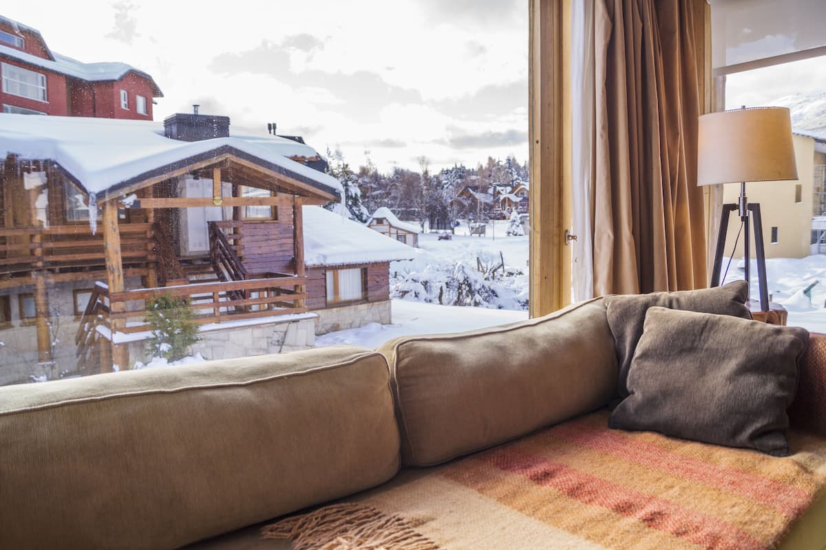 A cozy room with a view of a snowy village, a wooden cabin, and a lamp by the window.