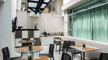 A modern dining area with glass tables and black chairs, a kitchen in the background, and a large window with curtains.
