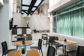 A modern dining area with glass tables and black chairs, a kitchen in the background, and a large window with curtains. - Hotel Asturias Medellin (Medellín)