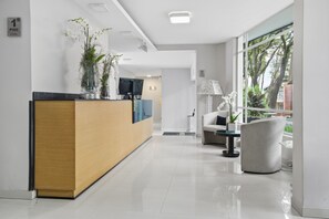 A modern reception area with a wooden reception desk, a flat-screen TV, and a seating area with a sofa and armchair. - Hotel Asturias Medellin (Medellín)