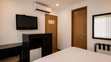 Ein Hotelzimmer mit einem Bett, einem an der Wand montierten Fernseher, einer Holztür und einem Fenster mit Jalousien.