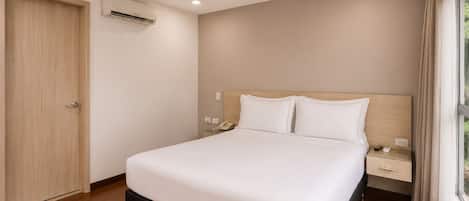 A hotel room with a bed, bedside tables, an air conditioning unit, and a window with curtains.