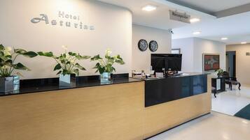A hotel reception desk with a sign reading 'Hotel Asturias,' a reception area with a flat-screen TV, a clock, and potted plants.