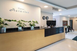 A hotel reception desk with a sign reading 'Hotel Asturias,' a reception area with a flat-screen TV, a clock, and potted plants. - Hotel Asturias Medellin (Medellín)